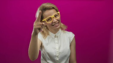 Sarı gözlüklü ve baş parmağıyla stüdyoda selam vermek için bekleyen parlak pembe duvarlı ve beyaz gömlekli bir kadın..