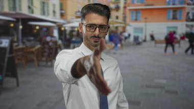 Beyaz gömlekli ve kravatlı adam, açık hava kafeleri ile sıralanmış sokakta el sıkışmak için elini uzatır; dostça selamlaşma.