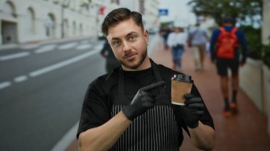 Önlüklü genç adam, şehir caddesinde beklerken kahve fincanına işaret ediyor. Açık havada eldivenli garson rolünün altını çiziyor..
