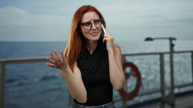 Sahil limanında telefonla konuşan, kızıl saçlı, gözlüklü ve siyah bluzlu bir kadın güneşli bir günde okyanusun arka planında konuşuyor.