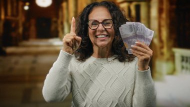 Kilisede elinde Rupiah banknotları tutan orta yaşlı bir kadın heyecanını dile getiriyor ve yukarıya doğru işaret ediyor. Mali güç ve kültürel bağlam tasvir ediyor..