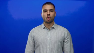 Gri çizgili gömlek giyen adam parlak mavi stüdyo arkaplanı önünde dudaklarını büzüyor, yakın plan portresi;.