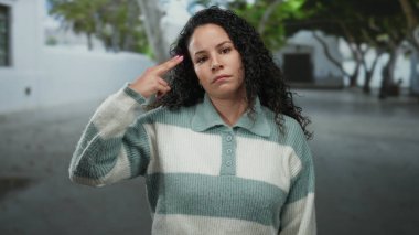 Kıvırcık saçlı bir kadın, sakin bir sokakta anlamlı jestler yapıyor. Duygularını, barışçıl bir arka plana karşı canlı bir şekilde sergiliyor..