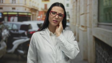 Bilim adamı üniformalı İspanyol kadın sokakta dikiliyor. Diş ağrısı ifadesi dışarıda. Etrafı arabalar ve eski binalarla çevrili..