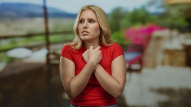 Gün boyunca açık hava restoranında titreyen, soğuk ve rahatsız görünen kırmızı gömlekli, manzara manzaralı, ısınmak için kendini kucaklayan, rahatsızlığını dile getiren bir kadın..