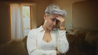 Beyaz tişörtlü, kısa saçlı bir kadın bina içinde el ele tutuşuyor, kapı ve perdenin önünde duruyor; utanç.