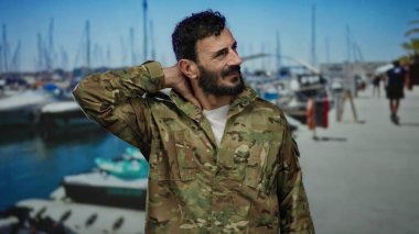 İspanyol üniformalı, sakallı bir adam tekneleri görünen hareketli bir limanda duruyor, parlak mavi gökyüzünün altında boynuna masaj yapıyor gibi görünüyor..