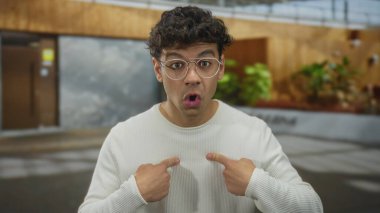 Dışarıdaki bina girişinde gözlüklü genç İspanyol adam parmak göğse işaret ediyor..