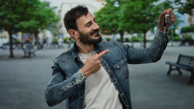 Sokakta elinde araba anahtarı olan İspanyol bir adam yeşil ağaçların altında kot ceket ve beyaz gömlek giyen neşeli bir gülümsemeyle işaret ediyor..