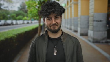 Arka planda ağaçlar ve arabalarla şehir sokağında el çırpan sakallı genç İspanyol bir adam sıradan moda ve doğal ifade sergiliyor..