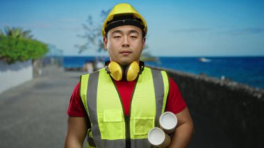 Elinde planlar tutan genç bir adam deniz kenarında sakin bir deniz altyapısı olan güvenlik ekipmanları giyiyor inşaat temaları ve açık havada mimari kavramlar sergiliyor..