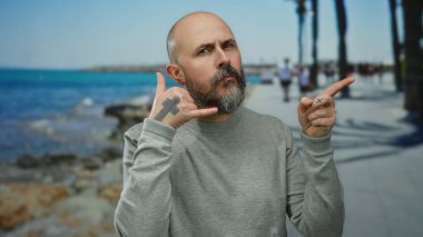 Kel sakallı adam deniz kenarında bir gezinti alanında telefon sembolünü işaret ederek arka planda rahatlamış bir açık hava manzarasını gösteriyor..