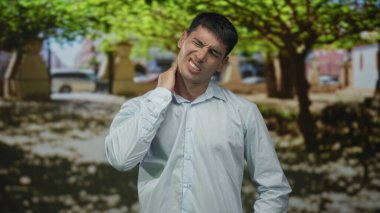 Mavi tişörtlü genç İspanyol adam açık havada güneşli yeşil parkta boynunu tutuyor..