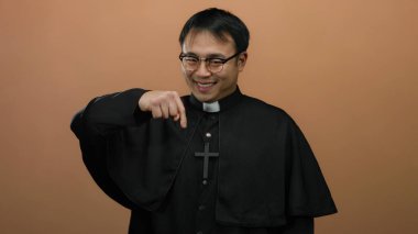 Kahverengi arka planda el kol hareketi yaparken gülümseyen ve işaret eden gözlüklü bir rahip, dostça bir ifade ve dini kıyafet sergiliyor.