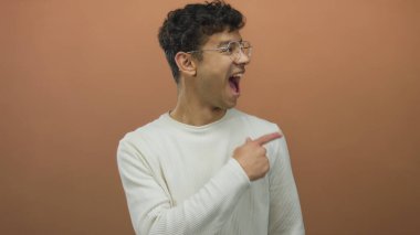 Adam beyaz süveter ve gözlük giyen turuncu stüdyo duvarına parmak işareti yapıyor; heyecan.