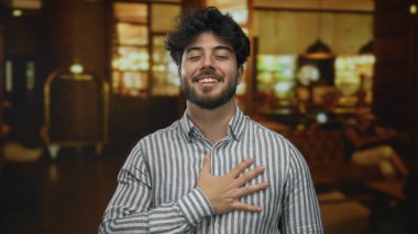 Otel lobisinde gülümseyen sakallı genç adam çizgili tişört giyiyor. Ilık, aydınlık, rahat bir atmosfer ve rahat bir iç mekan..