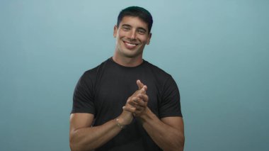 Stüdyoda gülümseyen ve alkışlayan İspanyol genç adam açık mavi arka plana karşı el çırpıyor..