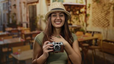 İspanyol kadın bir restoranın terasında gülümsüyor, elinde antika bir kamera, büyüleyici bir dekorla açık havada neşeli seyahat anları sergiliyor..