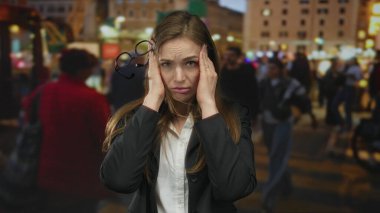 Gece vakti kalabalık bir aydınlık şehir sokağında gözlüğü yanında sallanırken, bir kadın şakaklarını iki eliyle bastırıyor..