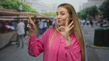 Pembe tişörtlü bir kadın şehir pazarında gülümseyip işaret ederken insanların ve tezgahların bulanık arkaplanlarını gösteriyor..