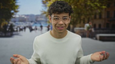 Beyaz kazaklı ve yuvarlak gözlüklü bir adam sokakta avuçlarını açık, kaşlarını kaldırmış ve başını hafifçe eğmiş olarak gösteriyor..