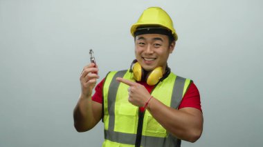 İnşaat malzemeleri ve beyaz arka planda sert bir şapka takan genç adam kendine güvenen bir gülümseme ve onaylanmış bir jest içeren bir cihaz sergiliyor.