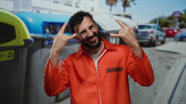 Turuncu hapishane tulumlu İspanyol sakallı adam şehir çöp konteynırının önünde kaya hareketi yapıyor. Gün ışığında yakalanmış..