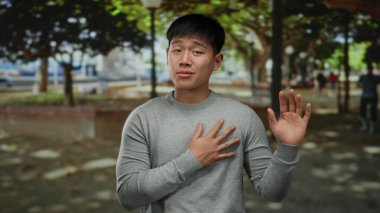 Genç Çinli bir adam, sakin bir açık hava parkında, gün ışığının altında el hareketleriyle duygularını ifade ederek iletişim için barışçıl ve çekici bir atmosfer yaratıyor..