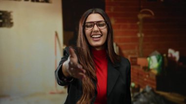 Gözlüklü İspanyol kadın gülümsüyor ve inşaat alanında elini uzatıyor. Kırmızı gömlek ve siyah ceket giyiyor. Arka planda aletler var..