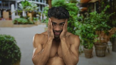 Bir şehir caddesi boyunca yeşil saksı bitkileri arasında başını iki eliyle kavramış çıplak bir adam; iç sıkıntı.