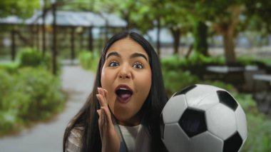 Yeşil park ortamında futbol topuyla tezahürat yapan, açık havada heyecan ve neşe ifade eden, futbol kültürü ve tutkusunu kucaklayan bir kadın..