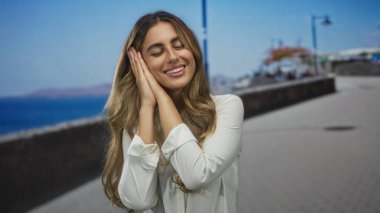 Genç sarışın kadın deniz kenarında bir dinlenme jestinde iki elini yanağına tutar; sakin rüya dinlenme.