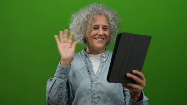 Gri saçlı, gülümseyen ve video görüşmesi sırasında yeşil arka planda tabletle el sallayan bir kadın..