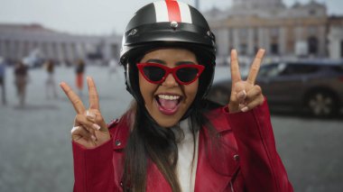 Aziz Peter Meydanı 'nda barış işareti ile poz veren kırmızı ceketli ve güneş gözlüklü bir kadın, kasklı tarzıyla Vatikan' ın çevresini canlı bir şekilde yakalıyor..