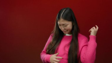 Genç bir kadın, izole edilmiş kırmızı bir arkaplan karşısında gitar hareketlerini şakacı bir şekilde taklit ediyor, etkileyici duygular ve canlı bir moda tarzı sergiliyor..