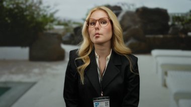 Otelin yanında yüzen, gözlüklü, siyah takım elbiseli, açık hava ortamında sarı saçlı bir kadın eğlenceli bir ortam yaratıyor..