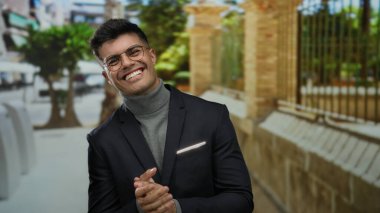 Gözlüklü genç İspanyol adam, bir şehir caddesinde gülümsüyor ve alkışlıyor, balıkçı yaka ve ceket giyiyor güneşli bir şehir zeminine karşı.