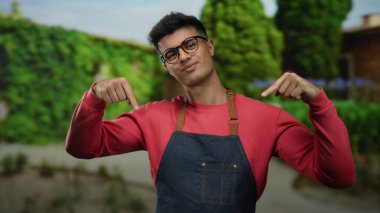 Gözlüklü ve önlüklü genç adam gülümsüyor ve yeşillik ve patikalarla çevrili bir açık hava parkına işaret ediyor..