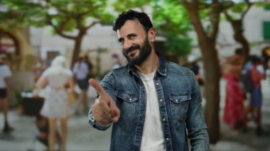 Sakallı İspanyol bir adam, arka planda bulanık insanlar ve ağaçlarla dolu kalabalık bir sokakta dikilirken 