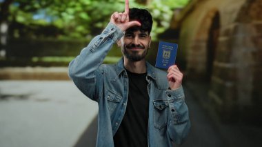 Dışarıda elinde İsrail pasaportu olan bir adam parmaklarıyla ezik jestler yapıyor, yeşil ve taş mimariyle çevrili kentsel sokak ortamında kot ceket giyiyor..