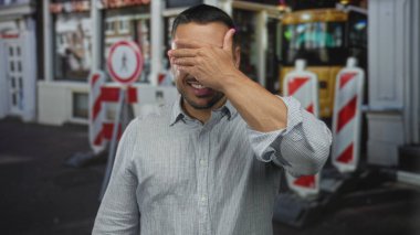 Kırmızı bariyerlerin yanındaki işlek caddede çizgili gömlekli genç adam yüzünü elleriyle kapatıyor..