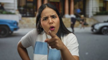 Genç Latin asıllı öfkeli konuşmacı kameraya bakarken kadın bir caddeyi işaret ederken parmağını uzatıyor..