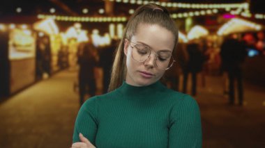 Yeşil kazaklı, yuvarlak gözlüklü ve kollu bir kadın bir gece fuarında parlak ışıklar altında çapraz; sessiz bir meydan okuma.