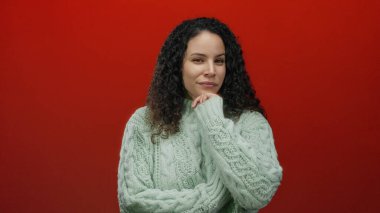 Yeşil kazaklı kadın canlı kırmızı bir arkaplanda düşünceli bir ifadeyle dikiliyor, kıvırcık saçlar sergiliyor ve izole bir arkaplanda sıradan bir moda sergiliyor..