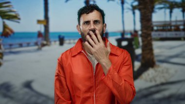 Turuncu hapishane tulumlu İspanyol bir adam sakallı güneşli bir sahil yürüyüşünde endişeli görünüyor, etrafta palmiye ağaçları var..