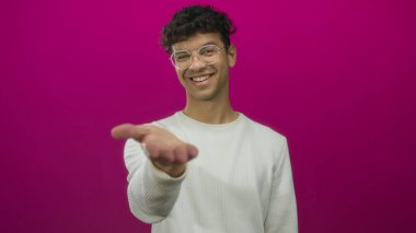 Kıvırcık saçlı, gözlüklü ve beyaz kazaklı bir adam pembe stüdyoda gülümserken elini açıyor; davet.