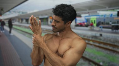 Tişörtsüz bir adam tren istasyonu inşaatı platformunda bileğini kavrarken tren arkasından süzülüyor..