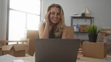 Genç bir kadın yeni oturma odasında yeni bir dizüstü bilgisayara bakıyor. Kutuları ve bitkileri taşıyor. Yeni bir başlangıç ya da ofis kurulumu öneriyor..