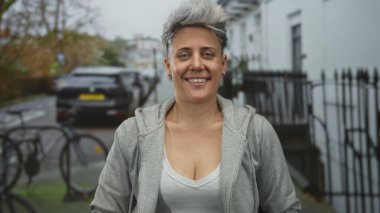 Gri kapüşonlu kısa gümüş saçlı kadın, bir şehir caddesinde gözleri kapalı gülümsüyor. Mutluluk..