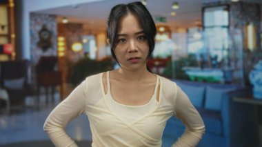 Bir kadın otel lobisinde durmuş modern dekorun ortasında ciddi bir ifade sergiliyor. Zarif bir ortamda sakin ve düşünceli bir ruh halini yansıtıyor..